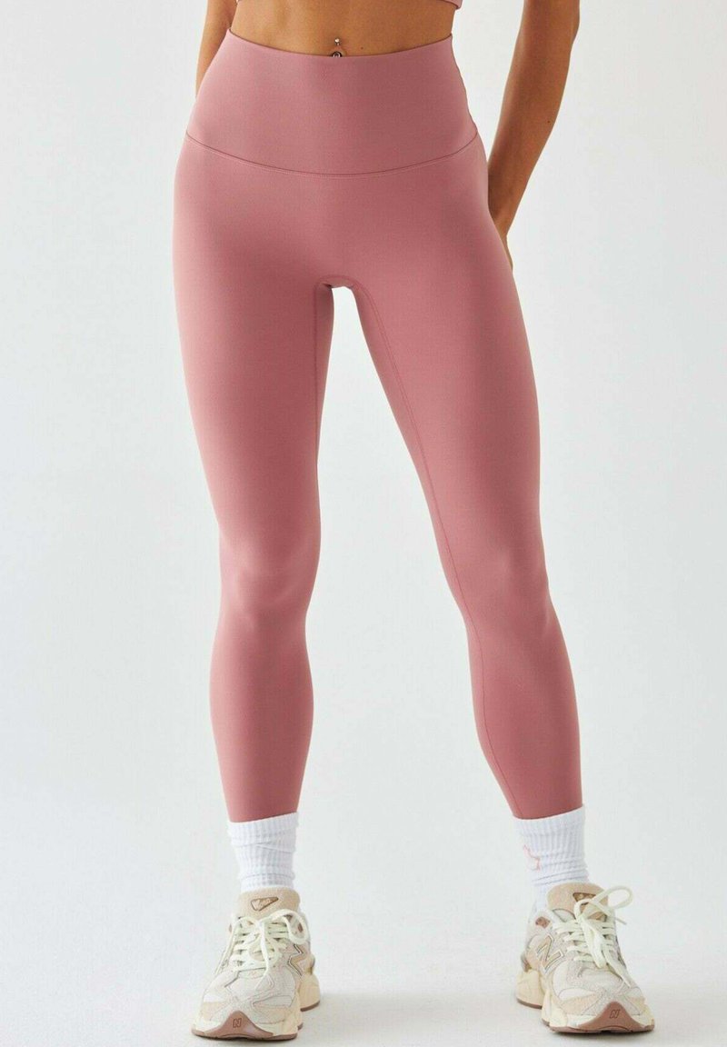 Różowe legginsy z wysokim stanem wykonane z elastycznego, gładkiego materiału, w połączeniu z beżowymi sneakersami i białymi skarpetkami za kostkę.