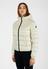 Giacca imbottita con cappuccio, chiusura con zip e polsini elasticizzati. Presenta una texture trapuntata e dettagli neri, indossata sopra pantaloni neri.