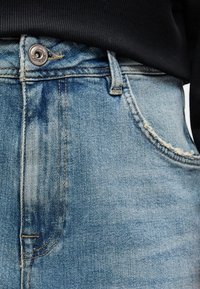 Ανοιχτό μπλε τζιν παντελόνι με κλασικό σχέδιο πέντε τσαντών, που διαθέτει ασημένιο κλείσιμο κουμπιού και ελαφριά φθορά γύρω από τις τσέπες.