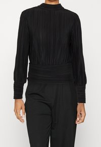 Blusa nera plissettata con maniche lunghe, colletto alto e una vita aderente. Il tessuto testurizzato con pieghe verticali crea un look strutturato.