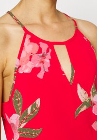 Top vermelho sem mangas com padrões florais em rosa e verde, apresentando um design de buraco na gola e tiras finas.