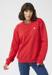 Red sweatshirt in a soft fabric, featuring a round neckline and ribbed cuffs, with a small white heart emblem on the chest.