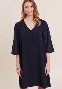 Marineblaues Kleid mit V-Ausschnitt, ¾-langen Spitzenärmeln und lockerer Passform. Glatte Textur mit einem subtilen Glanz im Spitzenmuster.