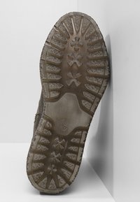 Semelle de chaussure en caoutchouc brun avec une surface texturée, présentant un motif d'adhérence large et une courbure vers le haut au niveau des orteils. Sale avec des traces d'usure visibles.
