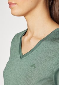 Groene V-hals T-shirt gemaakt van lichte, gestructureerde stof. Heeft een klein logo in een iets donkerdere schaduw op de linker borst.