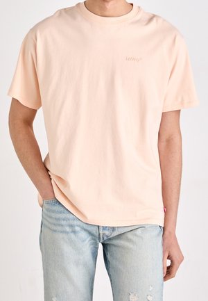 Mann trägt ein hellpfirsichfarbenes Levi’s T-Shirt mit kleinem Logo und hellblauen, abgenutzten Jeans, rechte Hand in der Hosentasche, steht vor weißem Hintergrund.