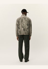 Jachetă gri, cu un model, cu guler înalt și mansete ribbate, purtată peste pantaloni verzi închisi. Cercul elastic și cusăturile subtile îi completează designul.