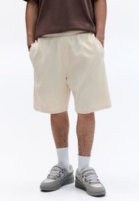 Beige Baumwoll-Shorts mit elastischem Bund, seitlichen Taschen und strukturiertem Stoff. Getragen mit grauen Turnschuhen und weißen Socken.