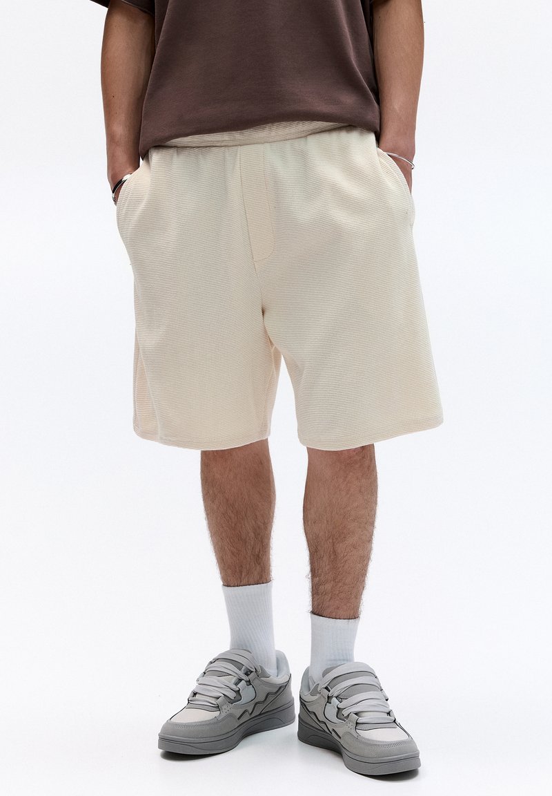 Beige Baumwoll-Shorts mit elastischem Bund, seitlichen Taschen und strukturiertem Stoff. Getragen mit grauen Turnschuhen und weißen Socken.
