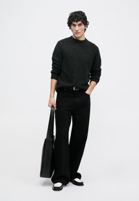 Black wool sweater, wide black pants, and black shoes with white accents. Holding a black tote bag, standing against a plain background.