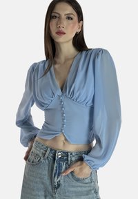 Hellblaue, kurz geschnittene Bluse aus transparentem Stoff mit V-Ausschnitt, Puffärmeln und Knopfleiste vorne. Kombiniert mit hoch taillierter Jeans.