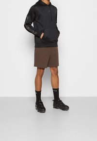 Svart hoodie med en framficka och logotyp, kombinerad med bruna shorts. Modellen bär svarta sneakers och strumpor med en vit logotypdetalj.