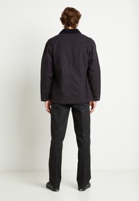 Giacca nera di cotone con colletto, taglio oversize e dettagli delle cuciture visibili. Abbinata a pantaloni neri slim e scarpe nere.