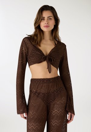 Donna che indossa un top corto marrone a maniche lunghe in crochet annodato sul davanti e pantaloni a vita alta coordinati in crochet con motivi a zigzag, in piedi su sfondo bianco.