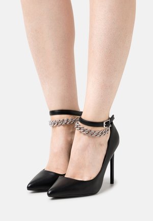 Escarpins noirs à talons hauts avec des bouts pointus, en cuir texturé, et une bride au cheville ornée d'un détail en chaîne argentée.