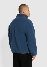 Giacca in pile di colore blu scuro con colletto alto, vestibilità rilassata e superficie testurizzata. Presenta polsini elasticizzati e orlo con cordino.