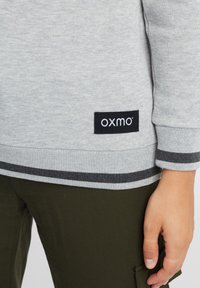 Felpa grigia chiara con polsini e orlo a costine, decorata con strisce nere e grigie, etichettata con un'etichetta nera "OXMO". Pantaloni verde oliva visibili.