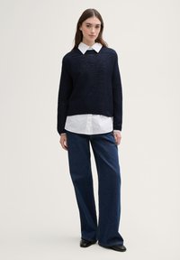 Pull en tricot bleu marine avec un motif texturé, associé à une chemise blanche à col et un jean denim à jambes larges. Des chaussures plates noires achèvent le look.