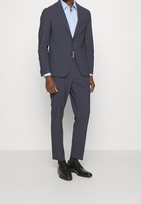 Man wearing a dark blue checked suit with black dress shoes and a light blue button-up shirt against a plain background.