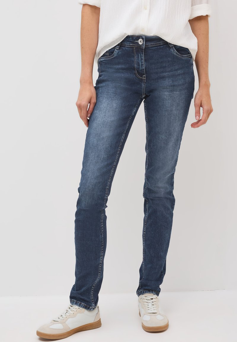 Jean en denim bleu avec une coupe slim, dotés de poches avant, d'une fermeture à bouton et de détails de coutures blanches, associés à des baskets blanches.