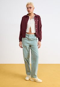 Casaco bomber bordô, top cropped canelado branco, calças verdes com riscas e sapatilhas bege; enquadrado num ambiente interior minimalista com chão amarelo.