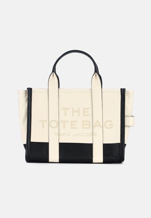 Beżowa skórzana torba typu tote z czarnymi akcentami i uchwytami. Na froncie znajduje się duży napis "THE TOTE BAG" oraz "MARC JACOBS".