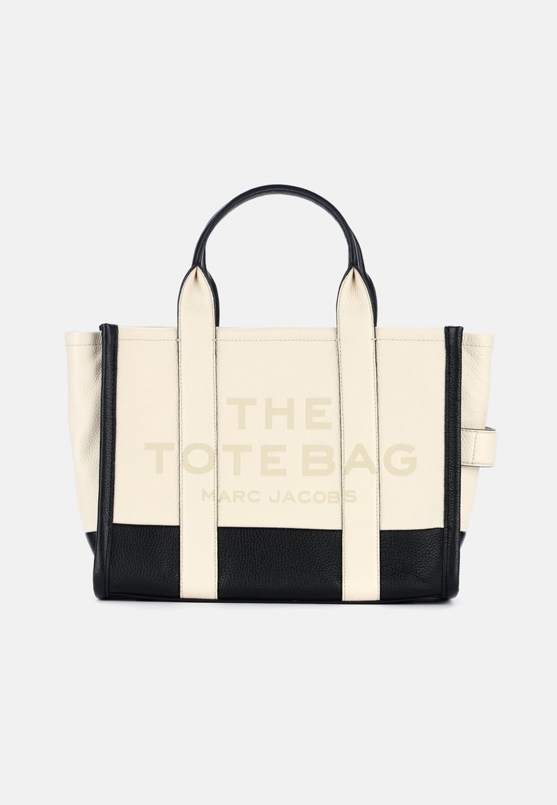 Bolsa tote de couro bege com detalhes e alças pretas. Apresenta um grande texto a dizer "A BOLSA TOTE" e "MARC JACOBS" na frente.
