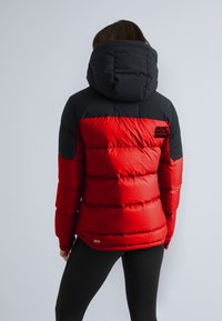 Rød og sort pufferjakke med hætte, der har en quiltet design, logodetalje på ryggen og elastiske manchetter. Bæres over sorte leggings.