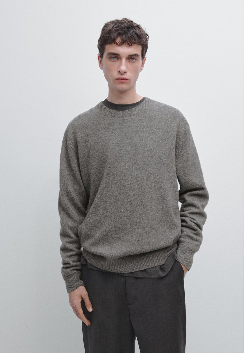 Pull en laine gris avec un col rond et des poignets côtelés, associé à un pantalon gris foncé. Le tissu semble doux et texturé.