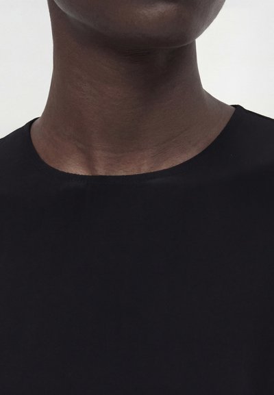 Κοντινό πλάνο του λαιμού και του πάνω μέρους του στήθους ενός ατόμου που φοράει μια απλή μαύρη μπλούζα με στρογγυλή λαιμόκοψη σε ένα απλό φόντο.