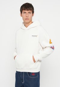 Sweatshirt branco com capuz e bolso frontal, apresentando o logotipo da McLaren e um detalhe amarelo em contraste na manga esquerda com a bandeira do Reino Unido.