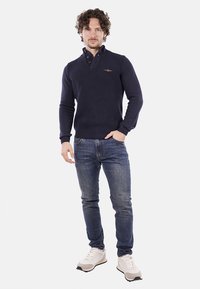 Maglione lavorato a maglia blu navy con colletto, chiusura a tre bottoni, indossato con jeans blu e sneakers bianche. Texture liscia e design aderente.