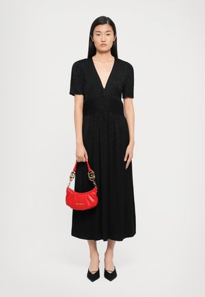 Frau mit langem, dunklem Haar, die ein schwarzes V-Ausschnitt-Midi-Kleid und schwarze High Heels trägt, hält eine kleine rote Handtasche mit goldenen Akzenten.