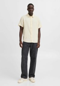 Kortärmad, ljusgul skjorta med knappar och struktur, kombinerad med mörka, löst sittande jeans och vita sneakers.