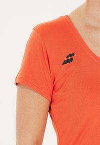 Nærbillede af en person iført en orange kortærmet skjorte med et sort geometrisk logo nær den højre skulder.