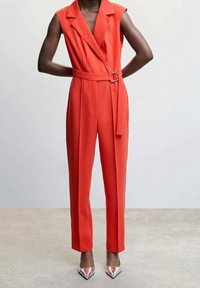 Czerwony kombinezon bez rękawów z paskiem w talii, o dopasowanym kroju i ostrym kołnierzu. Noszony z metalicznymi, spiczastymi szpilkami. Gładka faktura tkaniny.