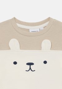 Beige ribbetopp med et kremfarget bjørneansikt-design; har broderte øyne, nese og to hvite ører på skuldrene.