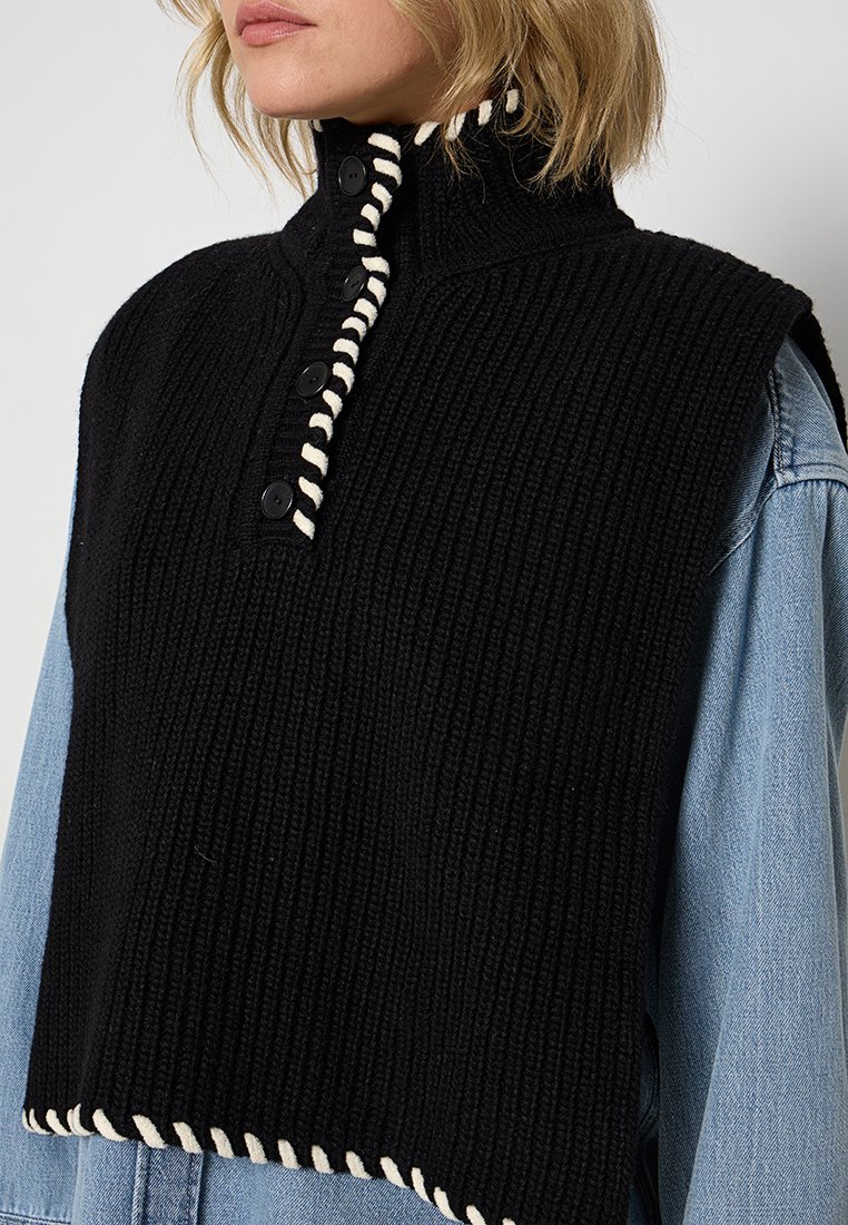 Schwarzes, gestricktes, ärmelloses Oberteil mit hohem Kragen, das über eine Knopfleiste und weiße Zickzack-Akzente entlang der Kanten verfügt. Strukturiertes Rippengewebe.