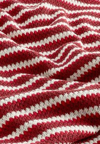 Strickdecke mit rotem und weißem Zickzackmuster, die eine strukturierte Oberfläche und drapierte Falten aufweist und ihr weiches, gewebtes Material betont.