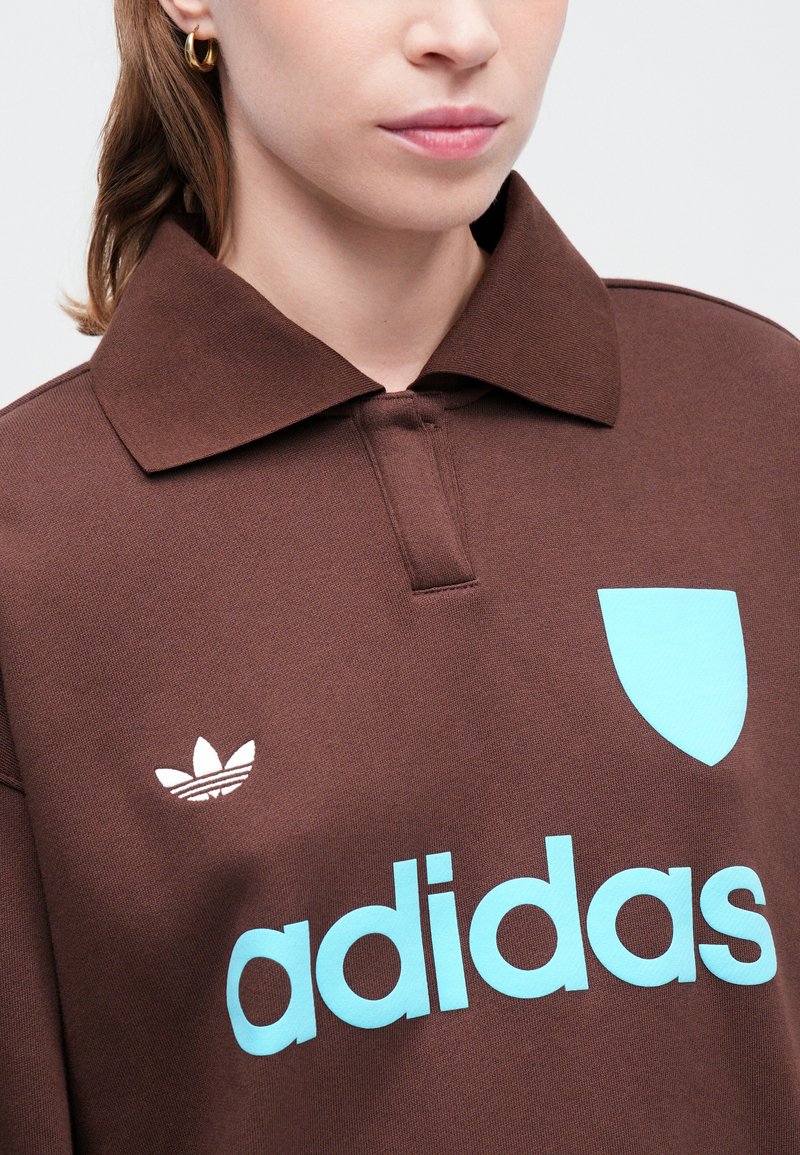 Polo shirt marron avec un col, présentant un logo et un design de blason bleu clair. Texture de tissu douce avec un look épuré et moderne.