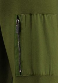 Tejido verde oliva con una textura suave; cuenta con un bolsillo con cremallera en el lateral para un almacenamiento seguro; líneas limpias y diseño simple.