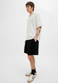 Hvid strikket polo med korte ærmer, prydet med et struktureret mønster, parret med sorte shorts og beige sneakers med farveaccentuer.