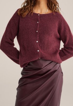 Frau trägt eine knopfgeöffnete, flauschige burgunderrote Strickjacke und einen passenden hochtaillierten, gerafften Lederrrock, steht mit den Händen in der Hüfte.