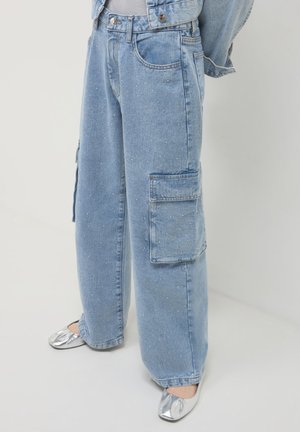 Helle blaue weit geschnittene Jeans mit kleinen weißen Punkten und einer seitlichen Cargo-Tasche, kombiniert mit glänzenden silbernen flachen Schuhen.