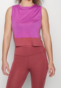 Nainen, joka on pukeutunut hihattomaan purppuran- ja vaaleanpunaisen sävyiseen urheilulliseen crop-toppiin ja korkeavyötäröisiin viininpunaisiin leggings-haalareihin, seisoo yksinkertaista taustaa vasten.