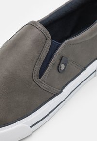 Chaussure gris à enfiler avec une tige en cuir lisse, des panneaux élastiques sur les côtés, et une semelle blanche avec une bande noire. Détails matériels minimaux visibles.
