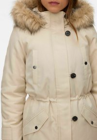 Bēšs parka stila mētelis ar kažokādas apdari pie kapuces, melnām pogām un elastīgu vidukli. Aprīkots ar divām sānu kabatām un gludu auduma faktūru.