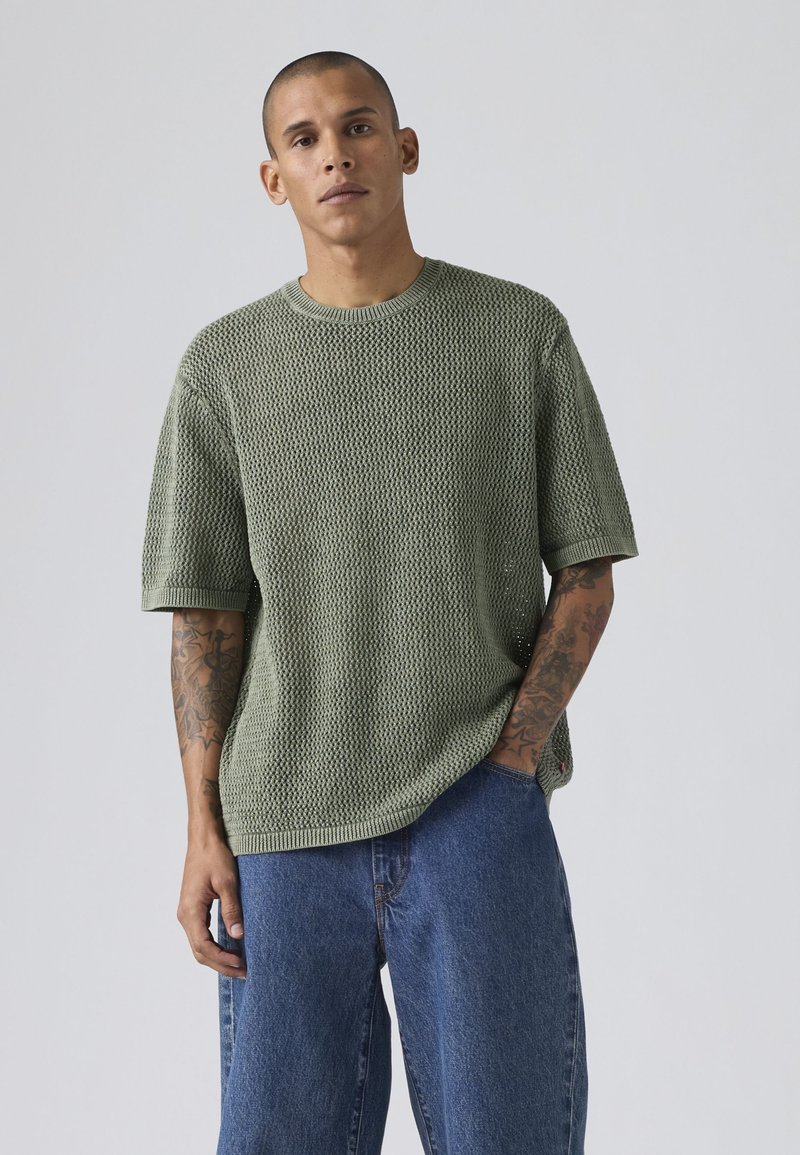 Hombre con brazos tatuados vestido con jersey verde texturizado de manga corta y vaqueros azules sueltos frente a un fondo liso y claro.