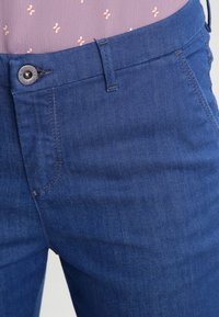 Close-up of blue denim jeans waist with metal button and pink shirt with small white floral pattern tucked in.
