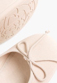 Ballet flats en toile beige avec un bout arrondi, un nœud décoratif et une semelle en caoutchouc à motifs floraux et circulaires.